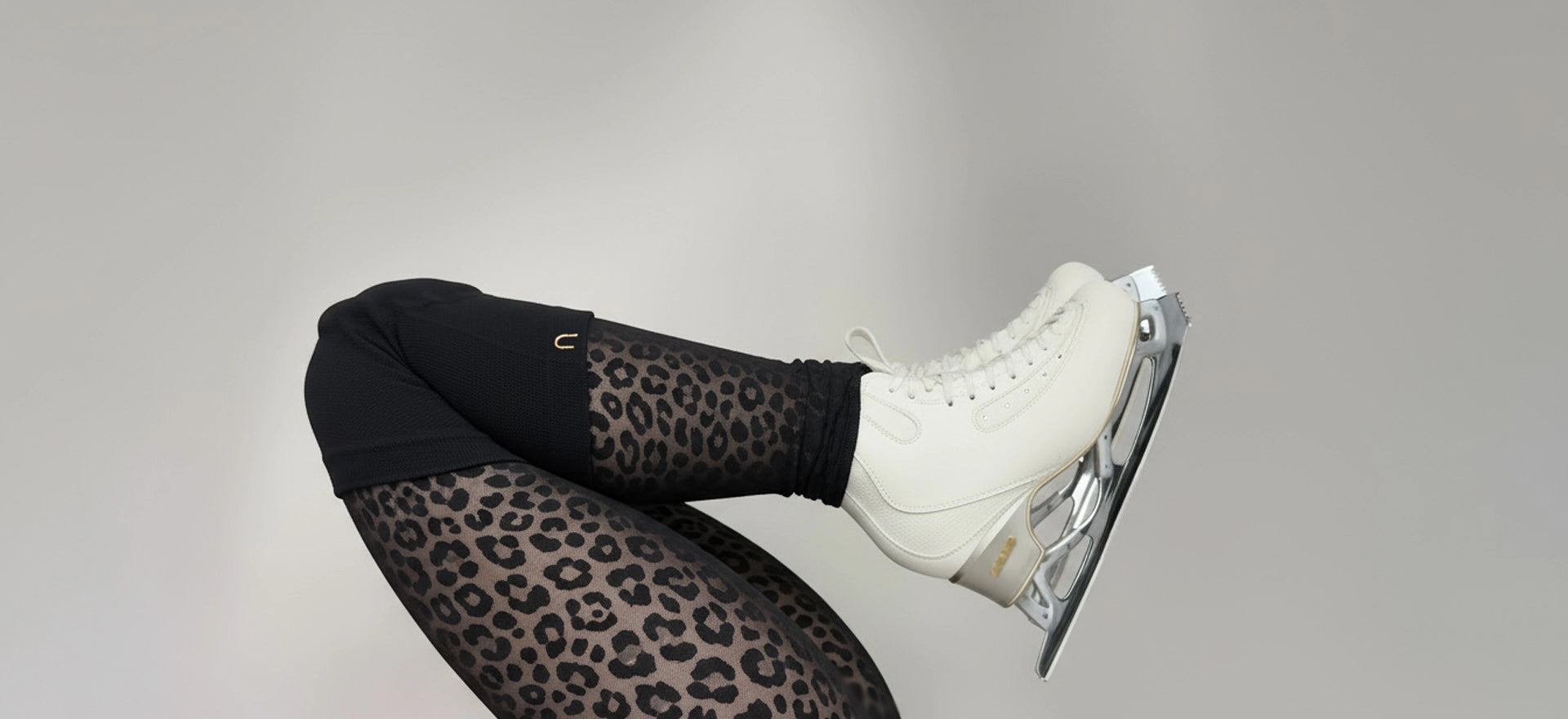 Figure skater wearing black knee guards and leopard print tights and edea ice skates on a gray background